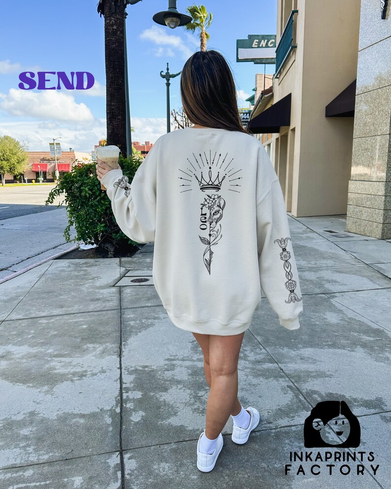 May include: A white sweatshirt with a black graphic print of a crown, a flower, and the word "OG" on the back. The sweatshirt has a black floral design on the sleeve.