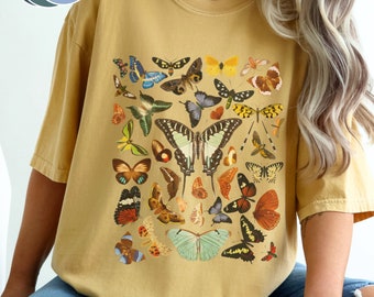 Puede incluir: Una camiseta amarilla mostaza con un estampado de mariposas de colores. Las mariposas están dispuestas en un grupo en el centro de la camiseta.