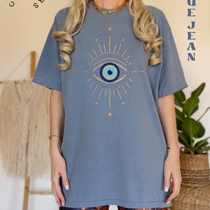 May include: A blue jean t-shirt with a gold evil eye design. The shirt is oversized and has a relaxed fit. The text "Comfort Colors" and "R" is visible in the top left corner of the image. The text "Blue Jean" is visible in the top right corner of the image.