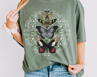 Puede incluir: Una camiseta verde con un diseño circular blanco que presenta una variedad de mariposas. Las mariposas son en tonos de marrón, negro, blanco y rojo. El texto "MOSS" está en la esquina superior derecha de la imagen.