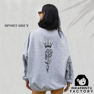 May include: A gray sweatshirt with a black graphic print of a crown, a flower, and the word "OOR" on the back. The sleeves have a black floral design. The sweatshirt is called "Sport Grey".