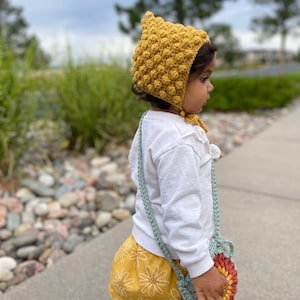 May include: A mustard yellow crochet bonnet with a textured, bobble stitch design. The bonnet is tied under the chin with matching yarn ties. A light blue crochet purse with a floral design is worn over the shoulder. The child is wearing a white long-sleeve shirt and yellow shorts.