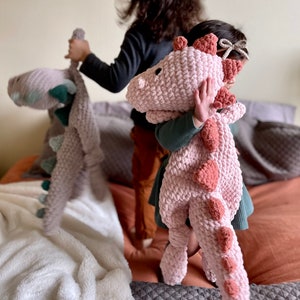 Pode incluir: Duas crianças brincando com brinquedos de dinossauro de crochê. Uma criança está segurando um dinossauro rosa com uma cauda longa, enquanto a outra criança está segurando um dinossauro cinza com detalhes verdes.