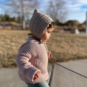Puede incluir: Un gorro de ganchillo beige claro con una parte superior puntiaguda y bordes festoneados. El gorro se ata debajo de la barbilla con una cuerda a juego. El niño lleva un suéter de punto acanalado rosa claro y leggings verde azulado con estampado floral.