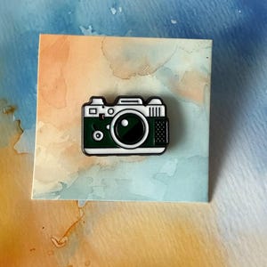 May include: A green and white enamel pin depicting a classic camera design. The pin has a black lens, a white body, and a black outline. It's placed on a watercolour background with blue, orange, and beige hues.