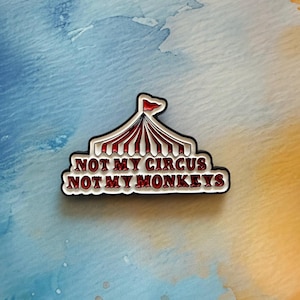 May include: A pin badge with a circus tent design in red and white stripes, topped with a red flag. The text "NOT MY CIRCUS NOT MY MONKEYS" is written in a stylized font, outlined in black.