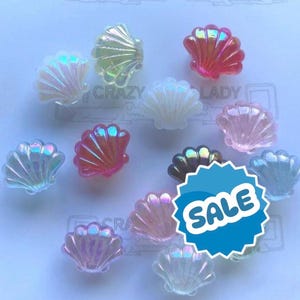 May include: A selection of iridescent seashell-shaped beads in shades of pink, white, and blue. The beads have a glossy finish and are arranged on a light blue surface. The word "SALE" is visible on a blue sticker.