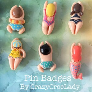 May include: Ten novelty pin badges depicting figures in swimwear. The badges feature various designs, including swimsuits, swim caps, and a mermaid tail. The colour scheme includes pink, yellow, blue, and brown, with the text "Pin Badges By CrazyCrocLady".