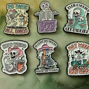 May include: A selection of enamel pins with humorous skeleton illustrations. Each pin displays a different slogan, such as "Even Baddies Get Saddies" and "Hot Mess Express." The pins are outlined in black and set against a green and yellow backdrop.