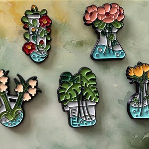May include: A selection of enamel pins with botanical themes. Each pin shows flowers or plants in a glass container, with a black outline and a teal border. The pins include red flowers, pink flowers, and green leaves.