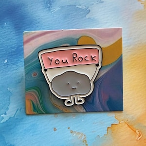 May include: An enamel pin with a smiling grey rock character holding a pink banner with the words "You Rock". The pin has a white border and is set against a colourful, abstract background. The pin is a fun, quirky accessory.
