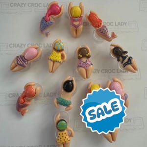 May include: A collection of eleven small, moulded plastic figures depicting swimmers in various swimsuits and poses. The figures feature diverse hair colours and bathing suit patterns, including polka dots and stripes. A blue star-shaped sticker reads SALE.