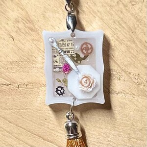 Puede incluir: Encanto de resina blanca con borla plateada. El encanto presenta una flor blanca, una llave plateada y un trozo de papel con la palabra "París" impresa.