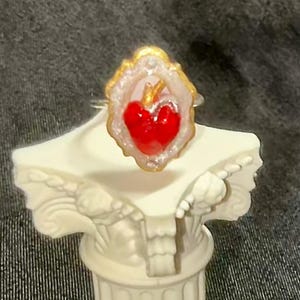 May include: A heart-shaped ring with a red center, framed by a white and gold border. The ring is set on a silver band. The ring is displayed on a white decorative column.