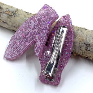 May include: A pair of pink glitter hair clips shaped like hands with white palmistry lines. The clips have silver metal clasps.