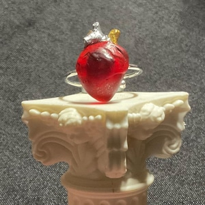 May include: A heart-shaped ring with a silver band. The heart is a translucent red with silver and gold accents at the top. The ring is displayed on a white decorative column.
