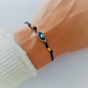 Puede incluir: Un brazalete de cuerda azul marino con un dije de ojo turco dorado. El dije tiene forma ovalada y tiene un ojo azul con una pupilla blanca.
