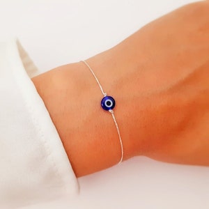 May include: A delicate silver chain bracelet with a small blue evil eye charm. The charm has a white center and is a popular symbol of protection.