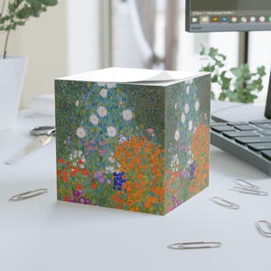 May include: A square paper memo cube featuring a vibrant floral print inspired by Gustav Klimt's garden paintings. The design displays a dense field of colorful flowers in shades of orange, red, purple, and white against a green background.