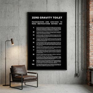 May include: A black framed poster with white text that reads "ZERO GRAVITY TOILET PASSENGERS ARE ADVISED TO READ INSTRUCTIONS BEFORE USE". The poster is mounted on a gray concrete wall. A brown leather chair and a black floor lamp are in the foreground.