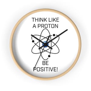 May include: A round wall clock with a light wood frame and a white face. The center features a black graphic of an atom with three small orbiting dots. The text reads THINK LIKE A PROTON BE POSITIVE! in black capital letters with black clock hands.