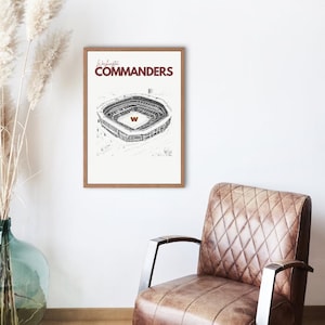 Puede incluir: Un cuadro enmarcado con una ilustración en blanco y negro de un estadio. El texto "Washington Commanders" está en borgoña sobre la imagen. La obra de arte está en un marco de madera, cerca de un sillón de cuero marrón.
