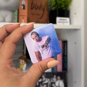 Puede incluir: Una chapa cuadrada con una impresi&oacute;n fotogr&aacute;fica de una persona con una camiseta rosa claro con texto oscuro, se&ntilde;alando hacia adelante con una mano. La chapa tiene un acabado brillante, esquinas redondeadas y un fondo azul y blanco.