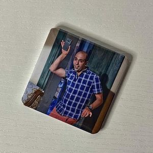 Puede incluir: Un posavasos cuadrado con la imagen impresa de una persona con la cabeza rapada que viste una camisa de manga corta a cuadros azules y blancos. La persona sostiene un peque&ntilde;o objeto rectangular oscuro hacia arriba con una mano mientras est&aacute; de pie.