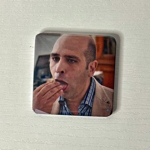 Puede incluir: Un im&aacute;n cuadrado con una impresi&oacute;n fotogr&aacute;fica de una persona calva de tez clara, que viste un blazer color canela sobre una camisa de botones a rayas azules y blancas, con la mano llevada a la boca como si estuviera comiendo o probando algo.