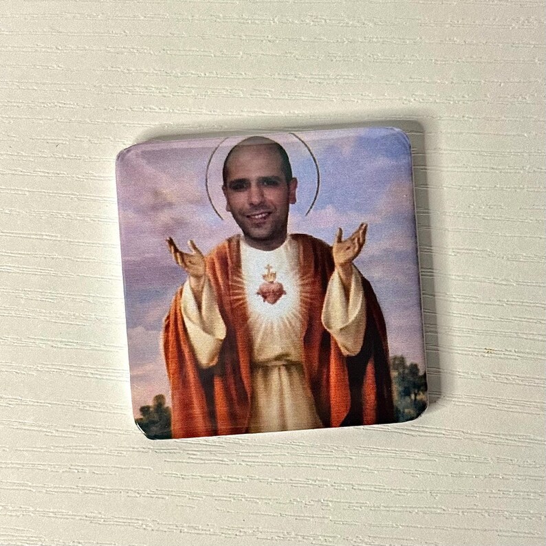 Puede incluir: Un im&aacute;n cuadrado con la imagen impresa de una persona con la cabeza rapada, representada al estilo de un icono religioso. La figura viste una t&uacute;nica beige con una capa roja, manos levantadas y un emblema de coraz&oacute;n radiante en el pecho.