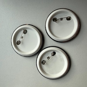 May include: Three circular pin-back buttons shown from the reverse side. Each button features a white plastic backing with a silver-toned metal pin mechanism attached horizontally across the center for fastening to fabric or other materials.