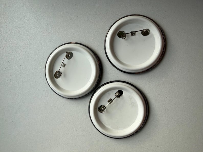 May include: Three circular pin-back buttons shown from the reverse side. Each button features a white plastic backing with a silver-toned metal pin mechanism attached horizontally across the center for fastening to fabric or other materials.