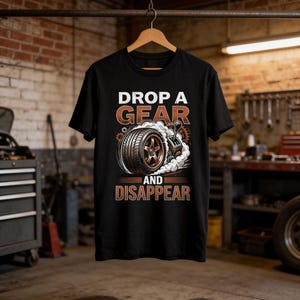 以下が含まれることがあります： 白とオレンジ色の「DROP A GEAR and DISAPPEAR」の文字が入った黒いTシャツ。グラフィックは、煙とギアシフトを備えた車のタイヤが特徴です。シャツは木製のハンガーに吊るされています。