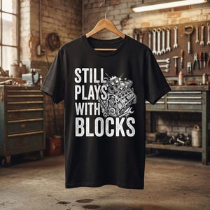 May include: Black t-shirt featuring the white text "STILL PLAYS WITH BLOCKS" and an engine graphic. The t-shirt is displayed on a wooden hanger. The background shows a workshop with tools and equipment, suggesting a mechanical theme.