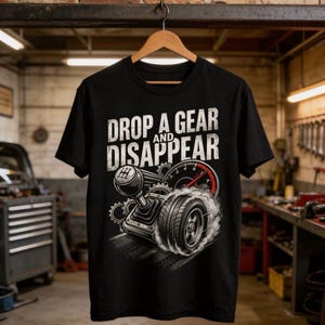 以下が含まれることがあります： 「DROP A GEAR AND DISAPPEAR」の白い文字がプリントされた黒いTシャツ。デザインには、ギアシフト、タイヤ、スピードメーターが含まれています。Tシャツは、木製のハンガーに吊るされ、ガレージに置かれています。