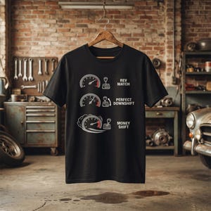 Puede incluir: Camiseta negra con un gráfico blanco de tres indicadores tipo velocímetro y texto. El texto dice "REV MATCH", "PERFECT DOWNSHIFT" y "MONEY SHIFT". La camiseta está colgada de una percha de madera.