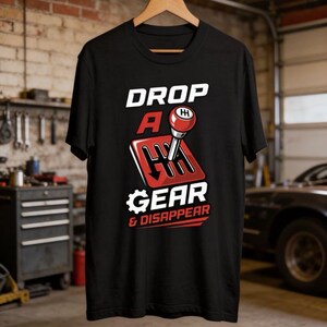 以下が含まれることがあります： 赤いギアシフトのグラフィックと白い文字「DROP A GEAR & DISAPPEAR」がプリントされた黒いTシャツ。木製のハンガーに掛けられており、ガレージの背景に配置されています。