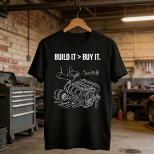 May include: Black t-shirt featuring the slogan "BUILD IT > BUY IT." and a white engine graphic with tools. The design shows detailed engine components, suitable for car enthusiasts and mechanics.