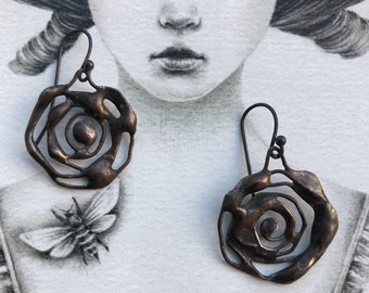 May include: A pair of bronze-colored earrings with a swirling rose design. The earrings are made of metal and have a delicate, intricate design.