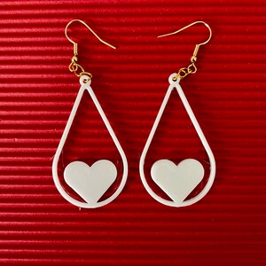May include: A pair of white teardrop-shaped earrings with a heart cutout. The earrings have gold hooks.