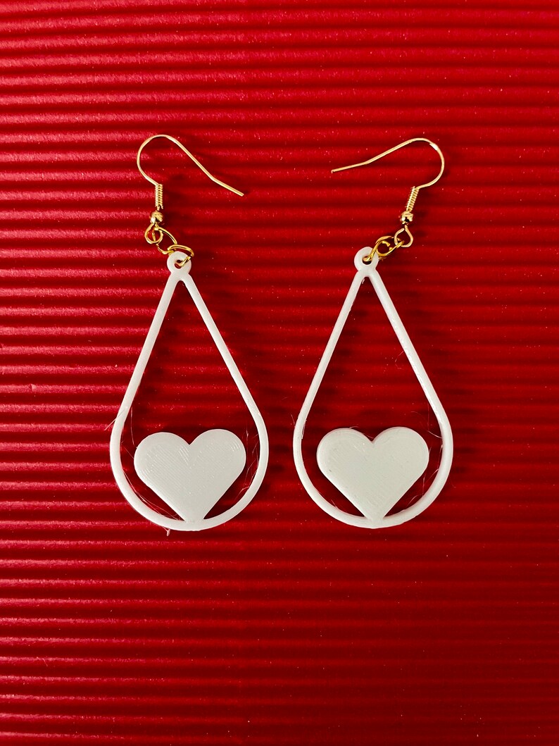 May include: A pair of white teardrop-shaped earrings with a heart cutout. The earrings have gold hooks.