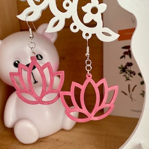 Puede incluir: Un par de pendientes de flor de loto rosa con ganchos plateados. Los pendientes cuelgan de un soporte de pendientes en forma de árbol blanco.