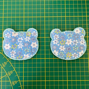 Pode incluir: Dois apliques de tecido azul claro em forma de ursos com um padrão floral branco. Os ursos estão em um tapete de corte verde.