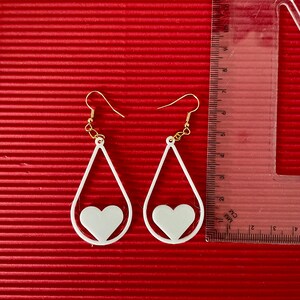 May include: A pair of white teardrop-shaped earrings with a smaller heart cutout inside each teardrop. The earrings have gold hooks.