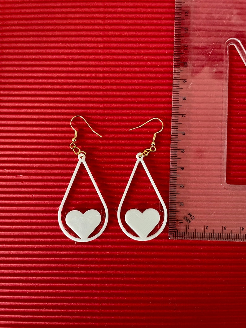 May include: A pair of white teardrop-shaped earrings with a smaller heart cutout inside each teardrop. The earrings have gold hooks.