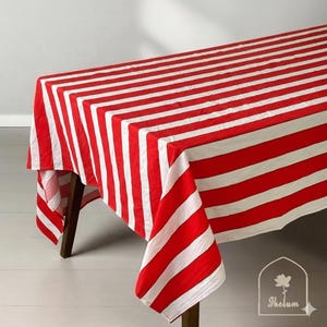 May include: A red and white striped tablecloth draped over a wooden table. The tablecloth features bold, evenly spaced stripes. The fabric is folded over the edges of the table, showcasing the pattern. The overall aesthetic is bright and clean.