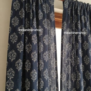 Peut inclure: Deux rideaux bleu foncé suspendus à une tringle en bois, ornés d'un motif répétitif d'arbres blancs et de fines rayures verticales. Le texte Indianlinenshop est imprimé deux fois sur le tissu. Les rideaux sont froncés en haut.