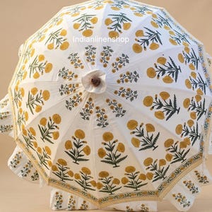 May include: A white parasol featuring a repeating floral pattern of yellow blossoms and green stems. The canopy has a ruffled edge with a thin yellow trim. The text Indianlinenshop is printed in the centre. The structure includes a brown finial.