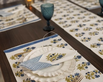 Puede incluir: Un camino de mesa blanco con un borde azul y un patrón floral en tonos verdes y azules. El camino de mesa está puesto con un plato blanco, una servilleta a rayas azul y blanca, y un tenedor y una cuchara de plata.