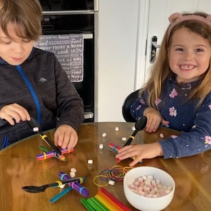 May include: Two children are seated at a wooden table, crafting with colourful craft sticks, rubber bands, and spoons. A bowl of marshmallows sits nearby. The children are focused on their projects, creating small airplane models.